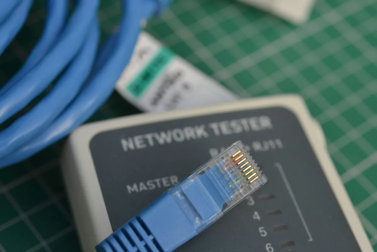 A blue Ethernet connector plugged into a handheld network tester on a green cutting mat, with coiled cable beside it, mid-diagnostic.